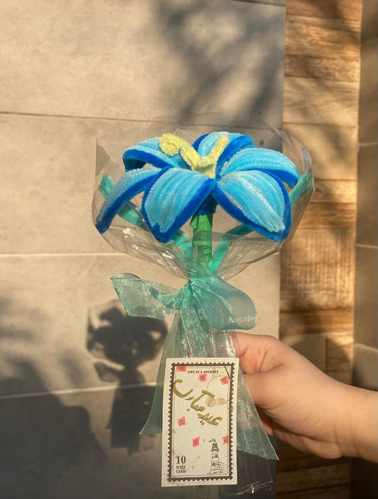 Blue and green decorative Small Lily Banquet wrapped in clear plastic with a hand holding it against a tiled wall.